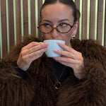Person wearing a brown fur coat holding a white cup against a striped wall.