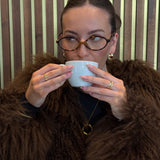 Person wearing a brown fur coat holding a white cup against a striped wall.