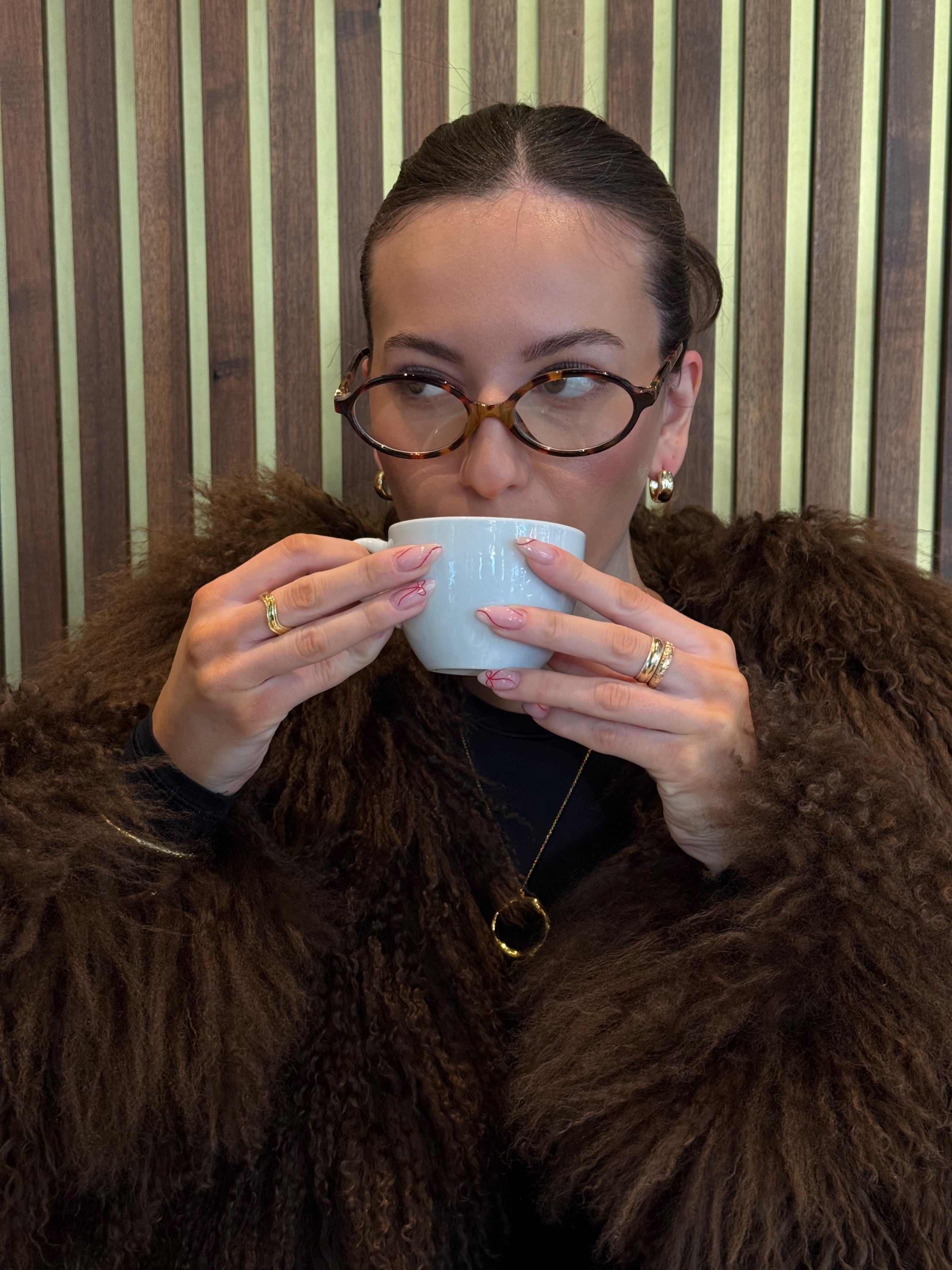 Person wearing a brown fur coat holding a white cup against a striped wall.