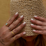 Person wearing a woven hat with hands covering the face, adorned with rings and nail polish.