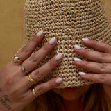 Person wearing a woven hat with hands covering the face, adorned with rings and nail polish.