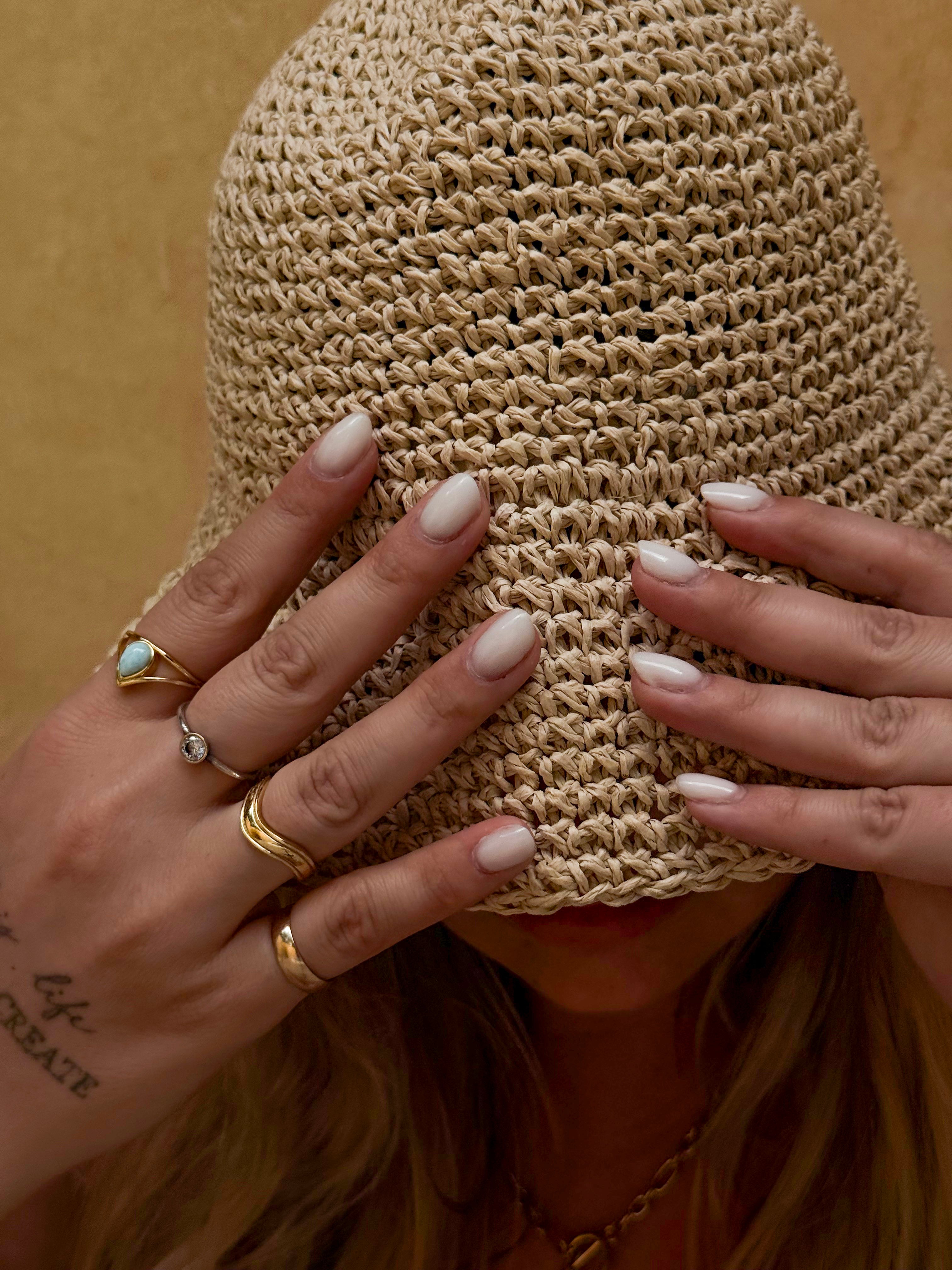 Person wearing a woven hat with hands covering the face, adorned with rings and nail polish.