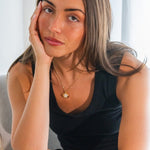 Woman wearing a black sleeveless top with a gold necklace, sitting against a light background.