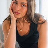 Woman wearing a black sleeveless top with a gold necklace, sitting against a light background.