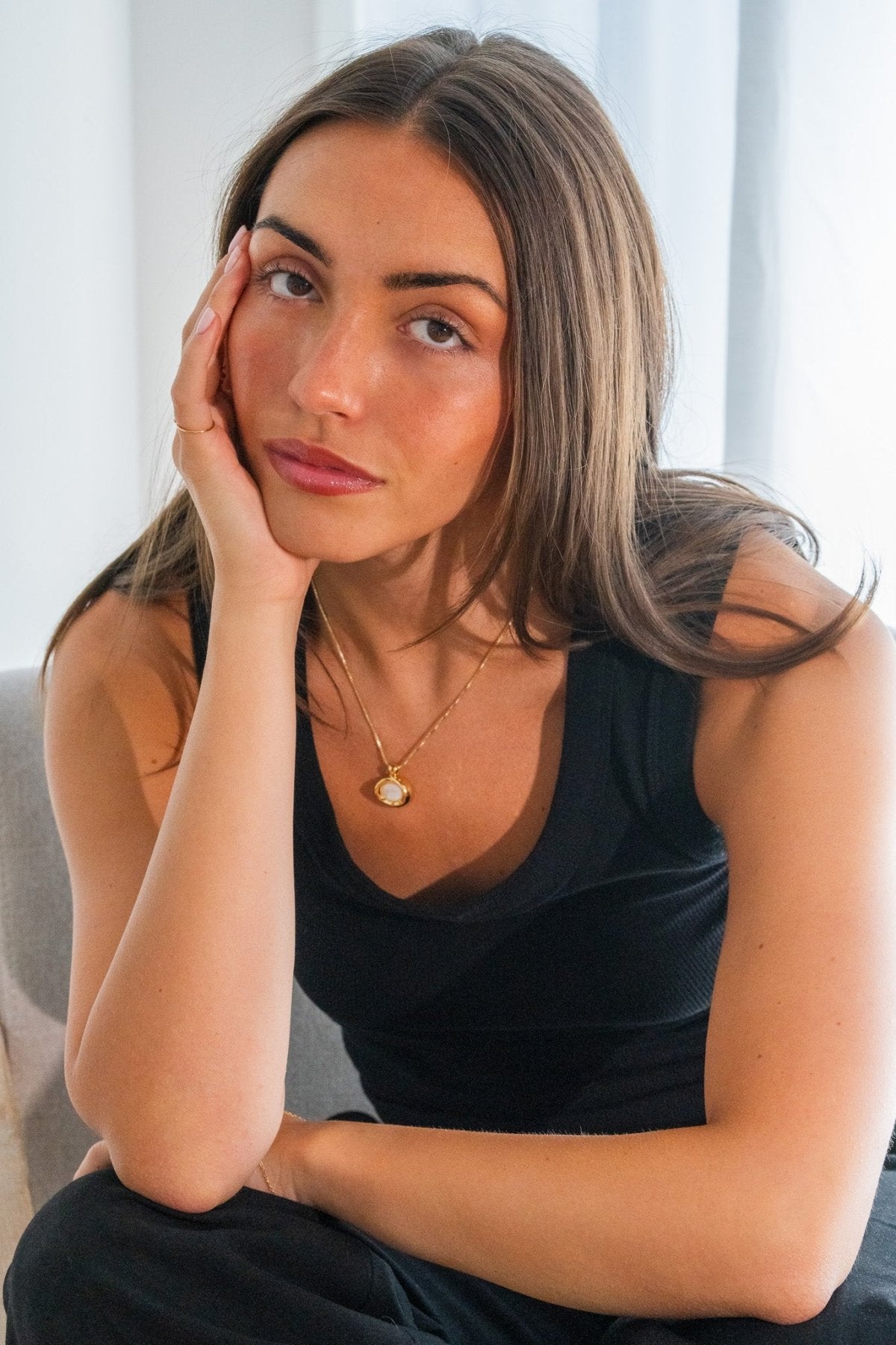 Woman wearing a black sleeveless top with a gold necklace, sitting against a light background.